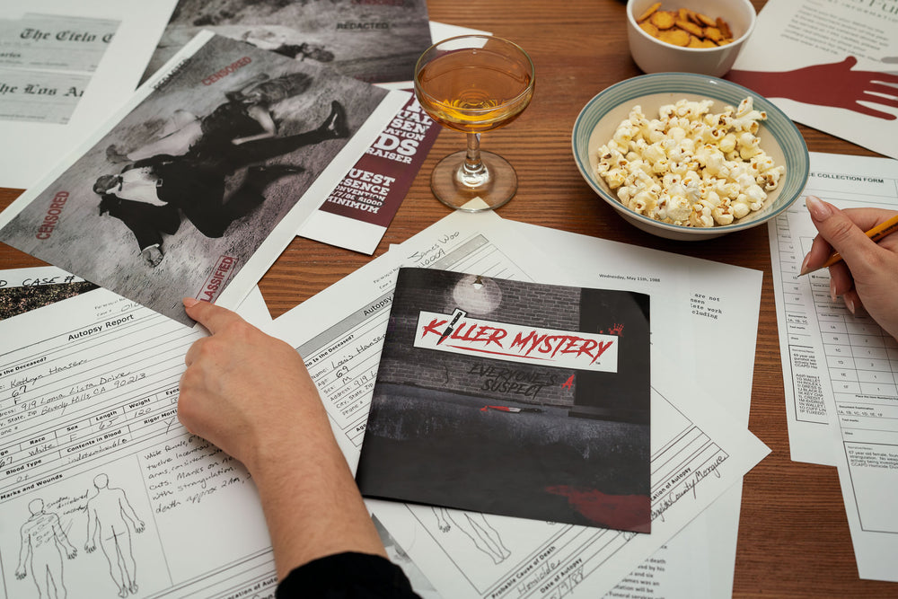 Picture of the contents of the game spread out onto a table, surrounded by snacks and drinks,  someone is pointing to one of the crime scene photos with one hand and has a pencil in the other hand to take notes.