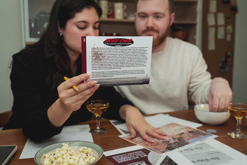 
            
                Load and play video in Gallery viewer, Pictured is a man and a woman sitting at a table playing the game. The woman is holding up the story card while the man reaches for some snacks. 
            
        